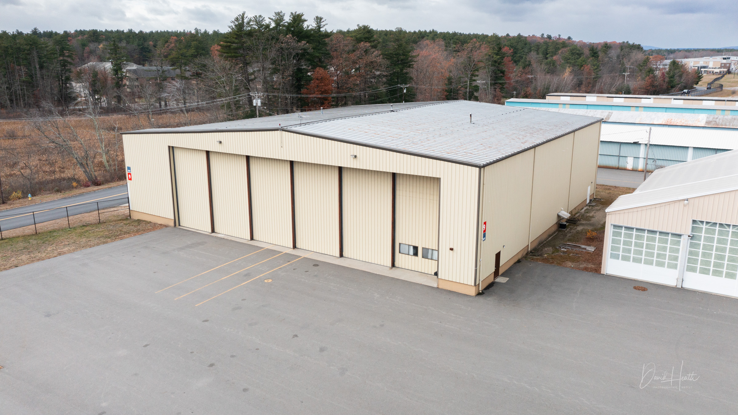 Aerial view of a beige industrial warehouse with large doors in a rural area, surrounded by trees and a paved lot.