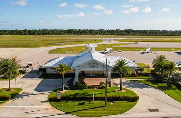 Small airport terminal with parked private jets, surrounded by palm trees and runways under a clear blue sky.