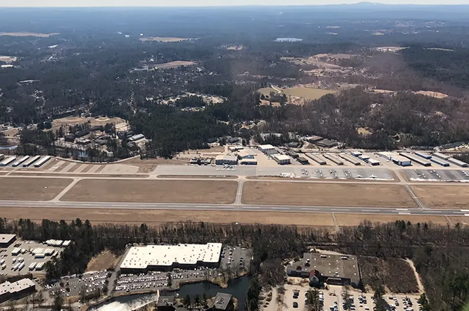 Aerial view of a rural airport with runways, parking, and surrounding forest landscape in daylight.
